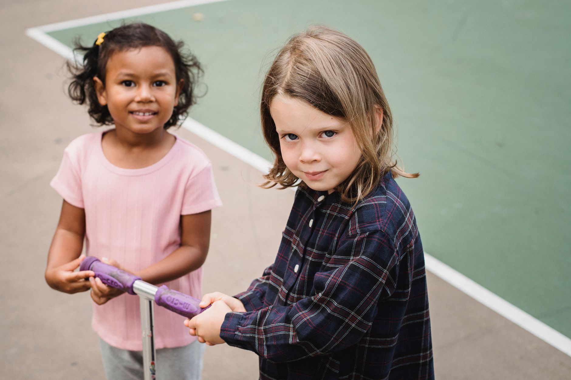 Two children, one with curly hair in a pink shirt and the other with long hair in a plaid shirt, are holding a purple scooter outdoors.